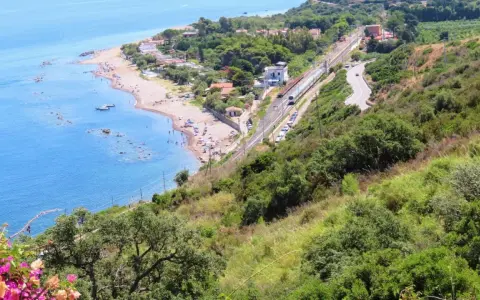 Spiaggia turchina finale di Pollina, con bagnanti e barche. Sopra, una ferrovia e una strada serpeggiano tra la vegetazione lussureggiante.