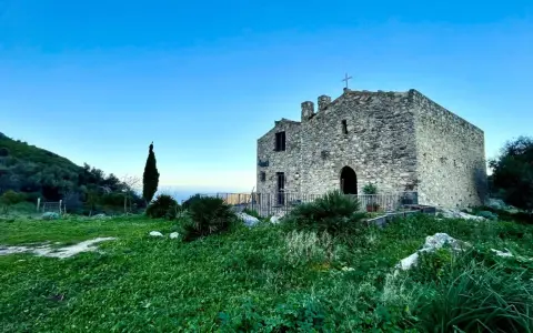 L'eremo di San Felice, un antico edificio in pietra con una croce sulla cima, si erge su una collina verdeggiante sotto un cielo azzurro.