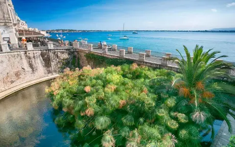 Siracusa, con la sua etimologia legata all'acqua, mostra papiri rigogliosi che galleggiano in un canale, con il mare azzurro e barche sullo sfondo.