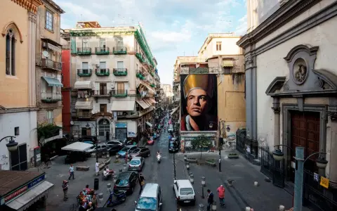 Un imponente murales di San Gennaro domina la scena in Via Forcella, Napoli. La strada è animata da auto, moto e persone.