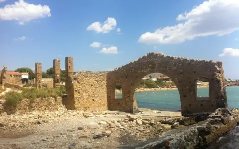 Rovine di una tonnara ad Avola, con un arco in pietra che si affaccia su una spiaggia affollata e il mare azzurro.