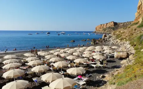 Spiaggia con ombrelloni e lettini, mare azzurro con barche e una scogliera. La torre conca domina il paesaggio.
