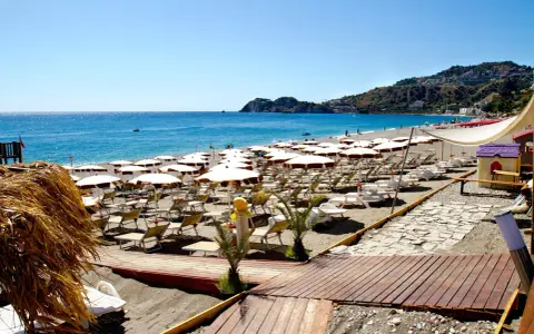 La spiaggia di Mazzeo, con file di ombrelloni bianchi e lettini, si estende verso il mare azzurro sotto un cielo limpido.