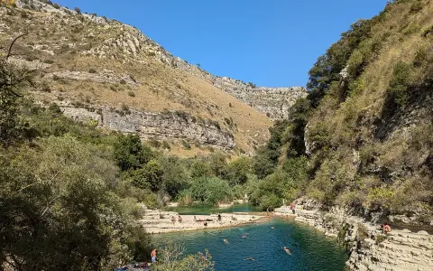 Gente si gode il sole e nuota nei laghetti d'Avola, circondati da rocce e vegetazione lussureggiante sotto un cielo azzurro.