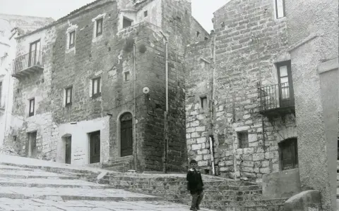 Un bambino cammina su una scalinata di pietra a Gibellina prima del terremoto. Edifici antichi in pietra la circondano.