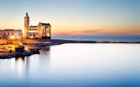 Tramonto sul porto di Trani, con la cattedrale illuminata che domina il lungomare. I vicoli del centro storico di Trani si intravedono tra gli edifici.