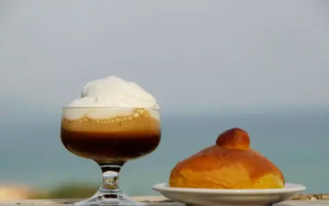 Un bicchiere di caffè freddo siciliano con panna e una brioche, con il mare sullo sfondo.