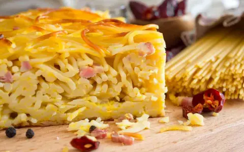 Fetta di spaghetti al forno, uno dei piatti tipici napoletani, con pancetta e formaggio. Sullo sfondo, spaghetti crudi e peperoncini.