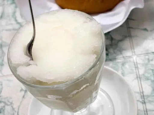 Un bicchiere colmo di granita al gelsomino, con un cucchiaino che affonda nella sua consistenza ghiacciata. Perfetta per una pausa rinfrescante.