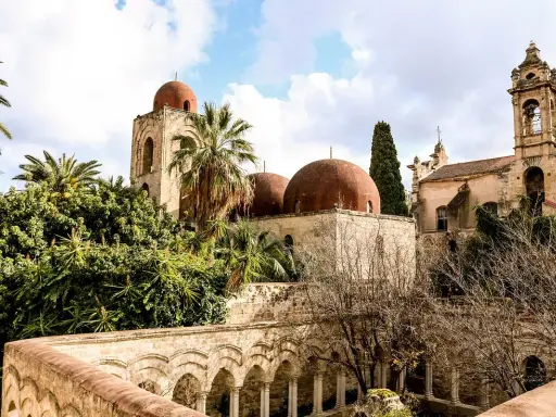 Le iconiche cupole rosse di Palermo si stagliano tra palme e vegetazione lussureggiante, un'antica bellezza sotto un cielo nuvoloso.