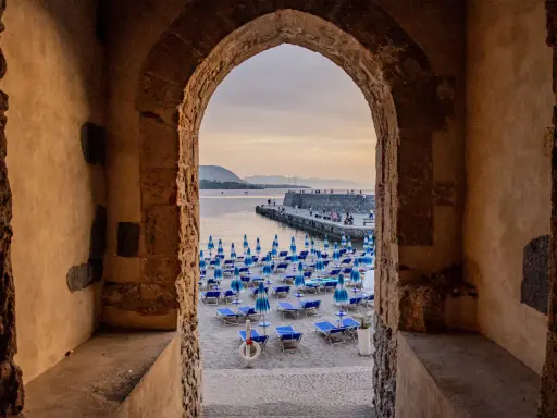 Arco antico incornicia la spiaggia di Cefalù con ombrelloni blu e lettini, vista sul mare e sul molo affollato.