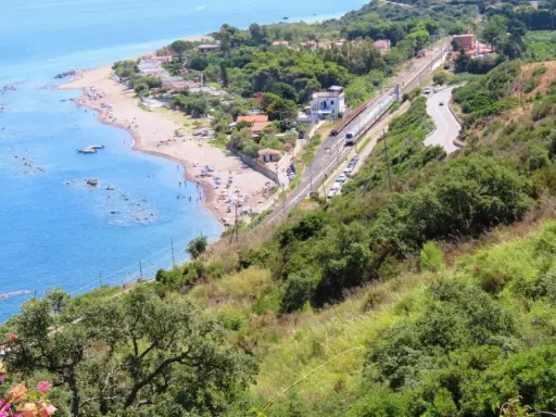 Spiaggia turchina finale di Pollina, con bagnanti e barche. Sopra, una ferrovia e una strada serpeggiano tra la vegetazione lussureggiante.