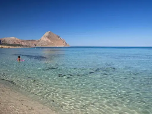 Bambina su un materassino rosa galleggia nelle acque cristalline della macari spiaggia, con montagne sullo sfondo.