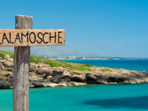 Cartello in legno "Calamosche Sicilia" su un palo, con mare turchese e costa rocciosa sullo sfondo.
