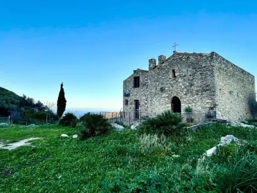 L'eremo di San Felice, un antico edificio in pietra con una croce sulla cima, si erge su una collina verdeggiante sotto un cielo azzurro.