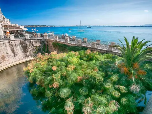 Siracusa, con la sua etimologia legata all'acqua, mostra papiri rigogliosi che galleggiano in un canale, con il mare azzurro e barche sullo sfondo.