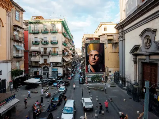 Un imponente murales di San Gennaro domina la scena in Via Forcella, Napoli. La strada è animata da auto, moto e persone.
