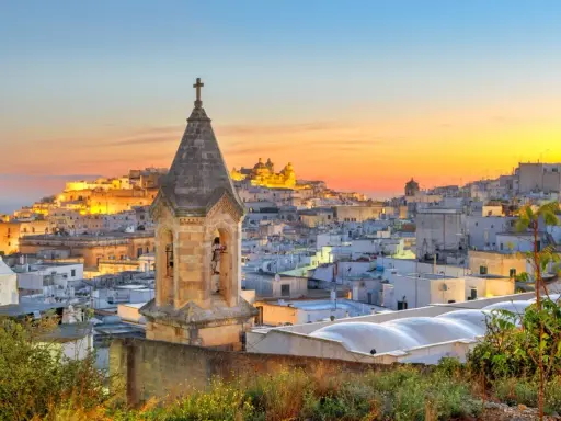 Campanile in primo piano, sullo sfondo la città bianca di Ostuni illuminata dal tramonto. Perfetto per scoprire cosa vedere a Ostuni in mezza giornata.