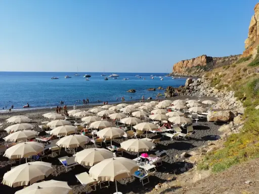 Spiaggia con ombrelloni e lettini, mare azzurro con barche e una scogliera. La torre conca domina il paesaggio.