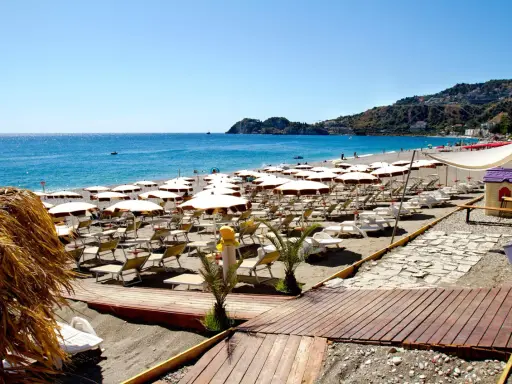 La spiaggia di Mazzeo, con file di ombrelloni bianchi e lettini, si estende verso il mare azzurro sotto un cielo limpido.