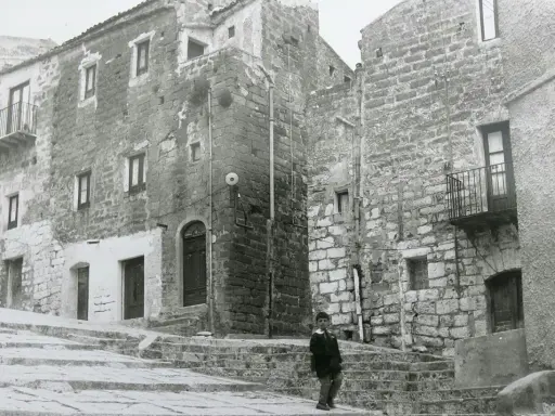 Un bambino cammina su una scalinata di pietra a Gibellina prima del terremoto. Edifici antichi in pietra la circondano.