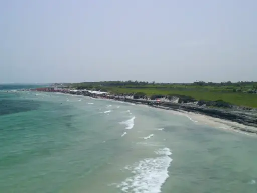 Vista aerea del lido Le Cesine, con acque turchesi e una spiaggia affollata di bagnanti e ombrelloni rossi.
