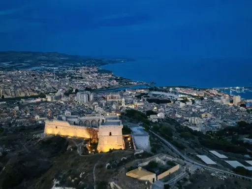 La rocca licata domina la città illuminata al crepuscolo, con il mare blu sullo sfondo.