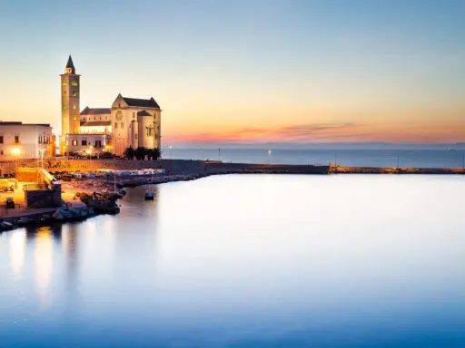Tramonto sul porto di Trani, con la cattedrale illuminata che domina il lungomare. I vicoli del centro storico di Trani si intravedono tra gli edifici.
