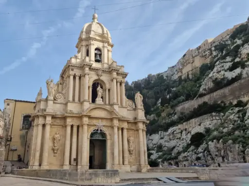 La chiesa barocca di San Giuseppe a Scicli, con il suo Cristo in gonnella, si erge maestosa contro una scogliera rocciosa.