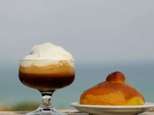 Un bicchiere di caffè freddo siciliano con panna e una brioche, con il mare sullo sfondo.