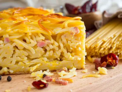 Fetta di spaghetti al forno, uno dei piatti tipici napoletani, con pancetta e formaggio. Sullo sfondo, spaghetti crudi e peperoncini.