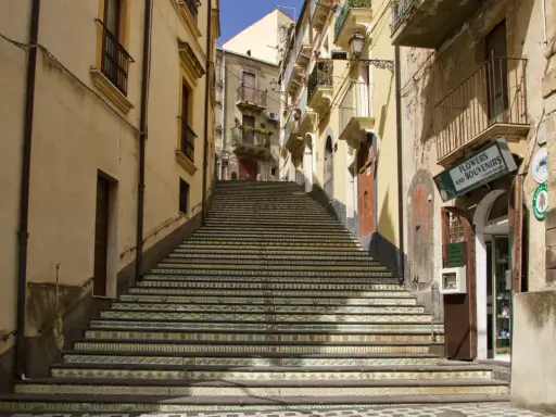 Una scalinata decorata con maioliche colorate sale tra edifici storici. Un negozio di fiori e souvenir invita a scoprire cosa vedere a Vizzini.