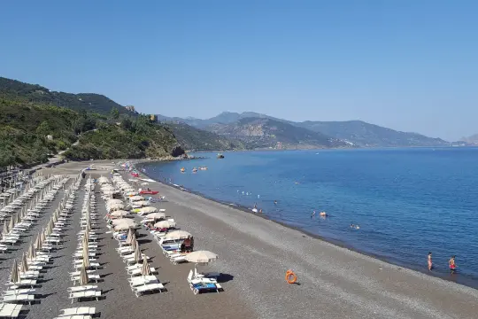 Spiaggia Torre Conca: file di lettini e ombrelloni su ghiaia scura, mare azzurro e colline verdi sullo sfondo.