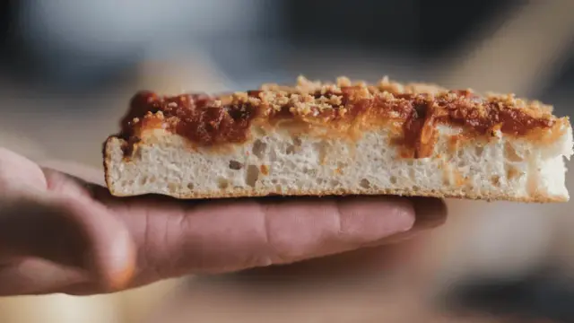 Una fetta di sfincionello palermitano, con la sua soffice mollica e il sugo rosso, tenuta in mano.