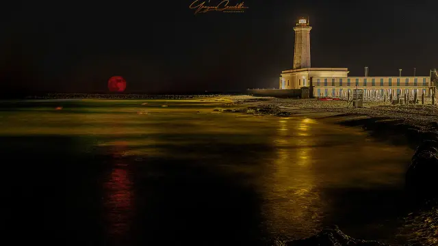 Un faro illumina il lungomare San Cataldo Lecce, mentre una luna rossa sorge sul mare calmo.