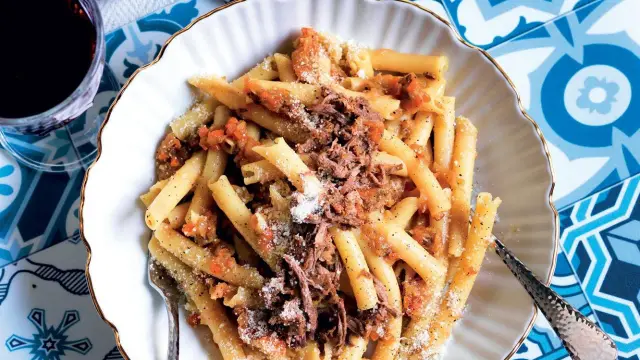 Piatto di pasta con ragù di carne, un classico dei piatti napoletani antichi, servito su un piatto bianco con bordo dorato, accanto a un bicchiere di vino rosso.