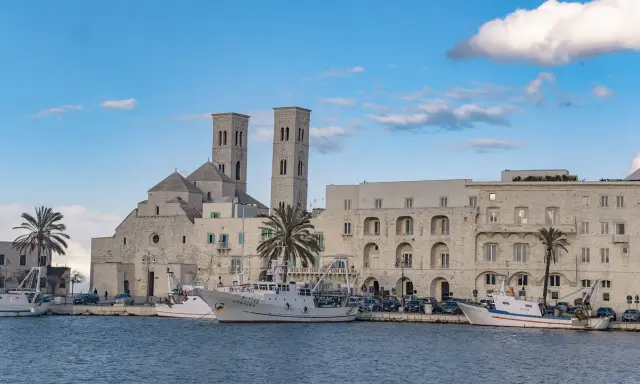 Molfetta cosa vedere: la Basilica e il porto con pescherecci sotto un cielo azzurro e nuvoloso.