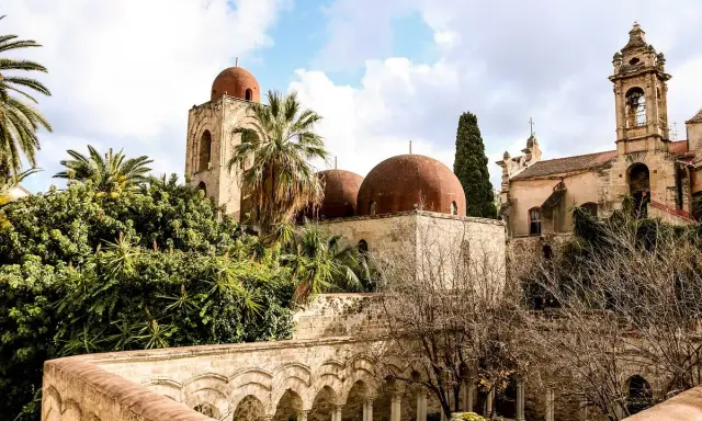 Le iconiche cupole rosse di Palermo si stagliano tra palme e vegetazione lussureggiante, un'antica bellezza sotto un cielo nuvoloso.