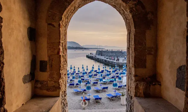 Arco antico incornicia la spiaggia di Cefalù con ombrelloni blu e lettini, vista sul mare e sul molo affollato.