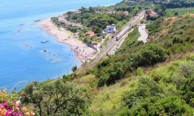 Spiaggia turchina finale di Pollina, con bagnanti e barche. Sopra, una ferrovia e una strada serpeggiano tra la vegetazione lussureggiante.