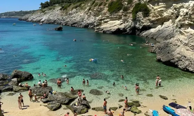Spiaggia 100 gradini Mongerbino, con acque cristalline e scogliere rocciose. Persone nuotano e prendono il sole in una giornata estiva.