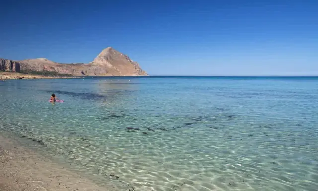 Bambina su un materassino rosa galleggia nelle acque cristalline della macari spiaggia, con montagne sullo sfondo.