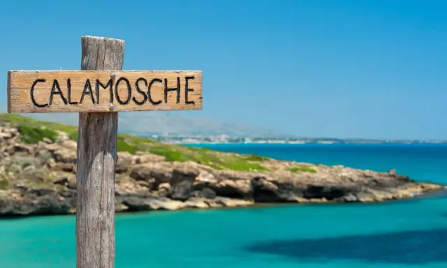 Cartello in legno "Calamosche Sicilia" su un palo, con mare turchese e costa rocciosa sullo sfondo.