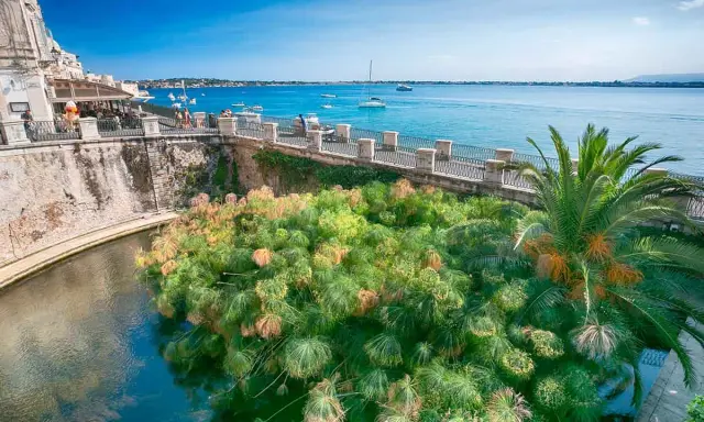 Siracusa, con la sua etimologia legata all'acqua, mostra papiri rigogliosi che galleggiano in un canale, con il mare azzurro e barche sullo sfondo.