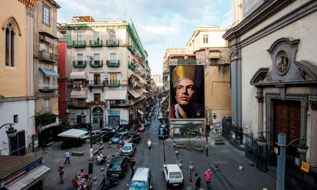 Un imponente murales di San Gennaro domina la scena in Via Forcella, Napoli. La strada è animata da auto, moto e persone.