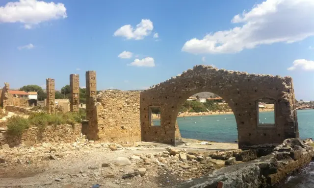 Rovine di una tonnara ad Avola, con un arco in pietra che si affaccia su una spiaggia affollata e il mare azzurro.