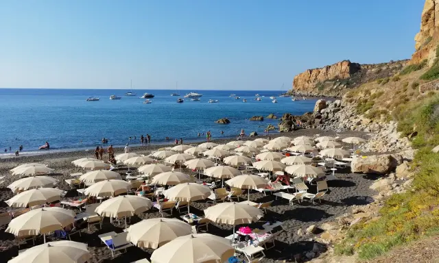 Spiaggia con ombrelloni e lettini, mare azzurro con barche e una scogliera. La torre conca domina il paesaggio.