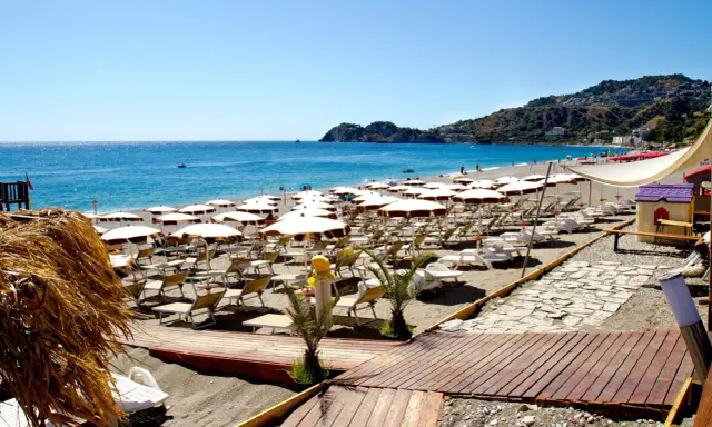 La spiaggia di Mazzeo, con file di ombrelloni bianchi e lettini, si estende verso il mare azzurro sotto un cielo limpido.