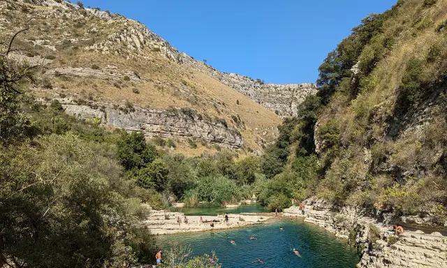 Gente si gode il sole e nuota nei laghetti d'Avola, circondati da rocce e vegetazione lussureggiante sotto un cielo azzurro.