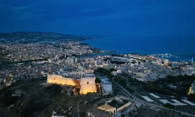 La rocca licata domina la città illuminata al crepuscolo, con il mare blu sullo sfondo.